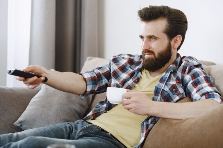 Homem no sofá aponta o controle remoto para a televisão enquanto segura uma xícara.